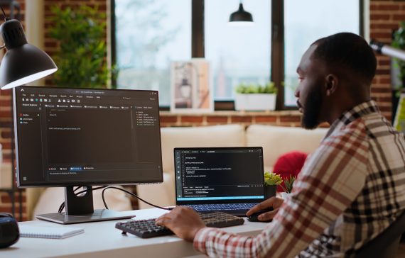 Male coder works from home typing code on a laptop in a modern living room. African american IT engineer using programming language and cloud computing for software development. Camera B.